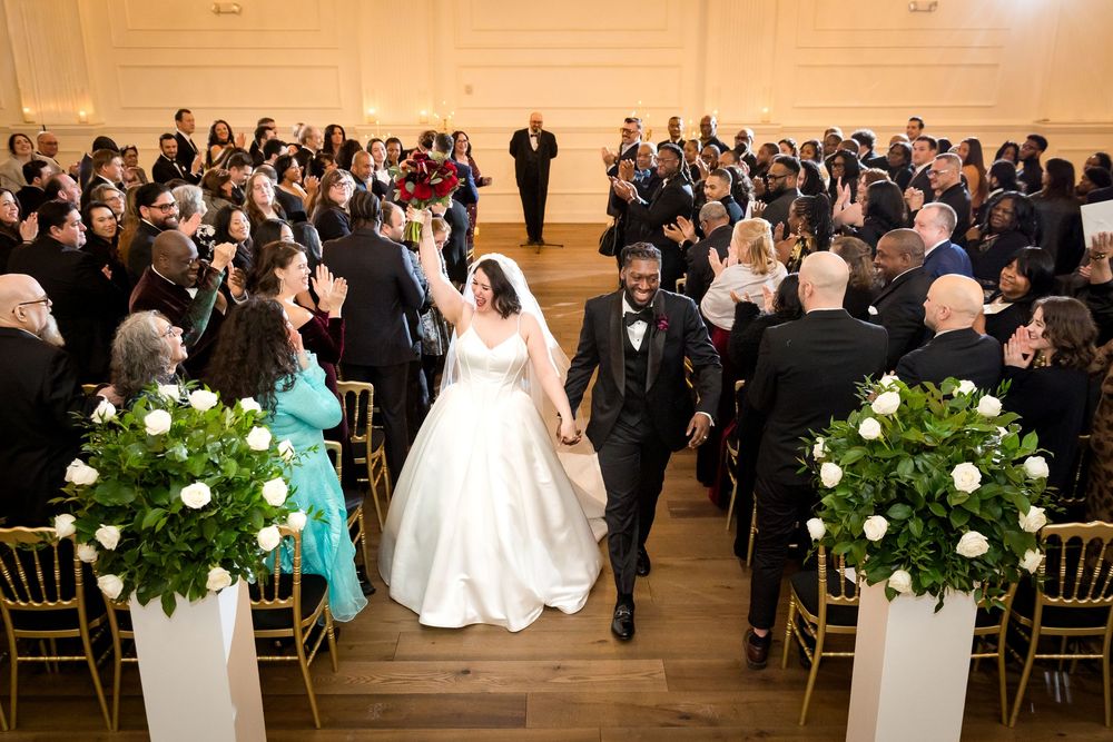 Bride and groom walk down the aisle holding hands after wedding ceremony in elegant white ballroom.