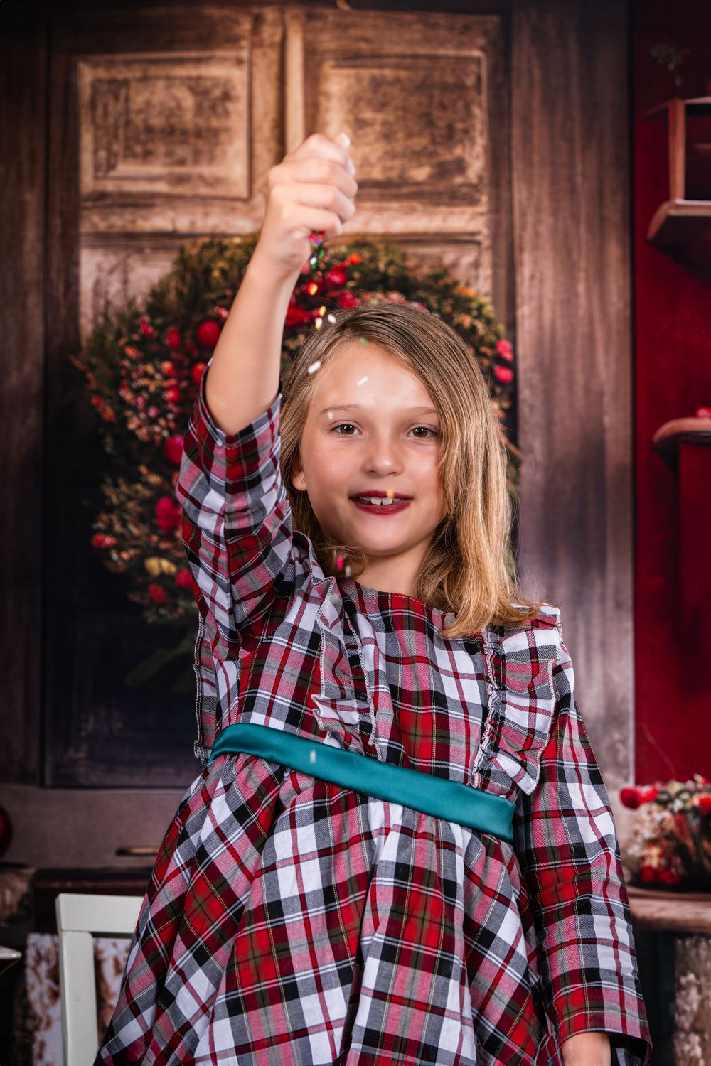 A young person in a plaid dress raises their arm triumphantly against a holiday backdrop.