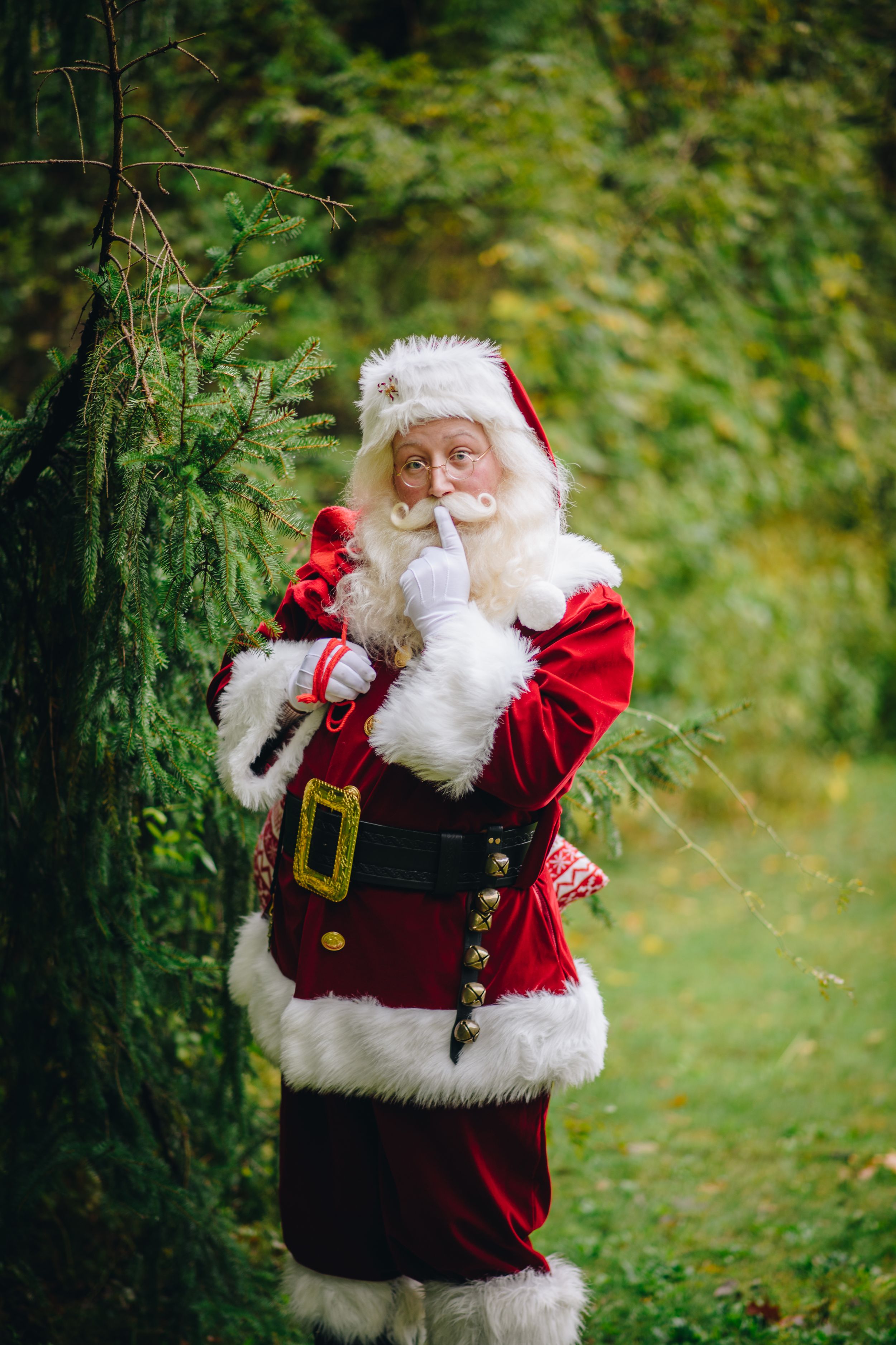 A figure in a traditional red and white Santa Claus costume stands in a garden making a shushing gesture.