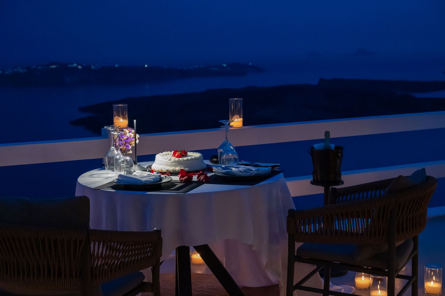 Six romantic tables illuminated by candlelight and string lights overlook a moonlit waterfront dining terrace at dusk.