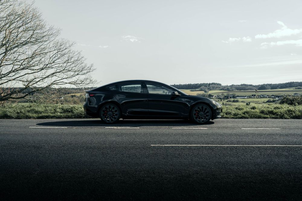 Side profile of a black Tesla Model 3 parked on rural road with countryside backdrop.