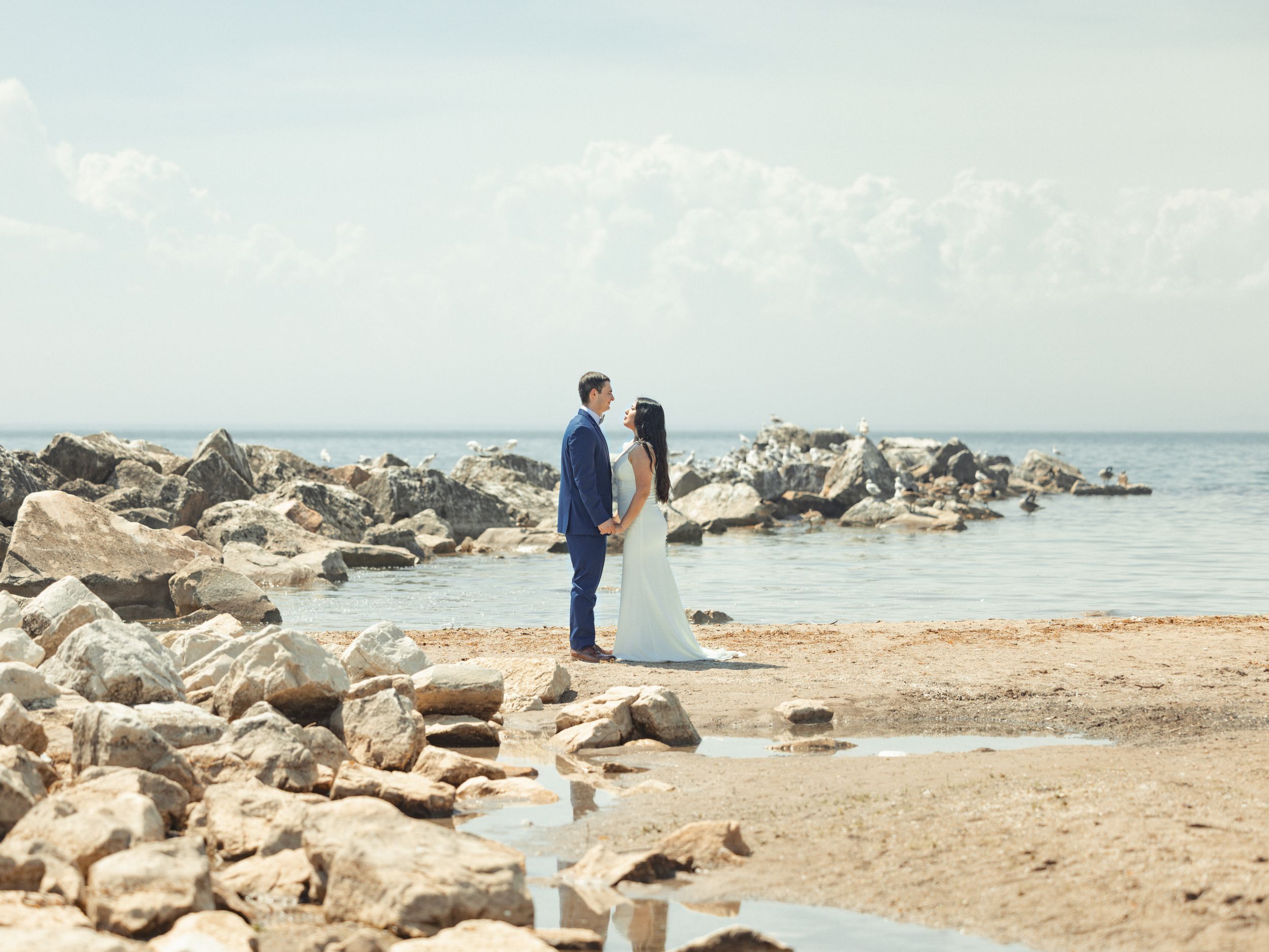 A couple stands facing each other on rocky coastal shore overlooking tranquil blue ocean and sky.