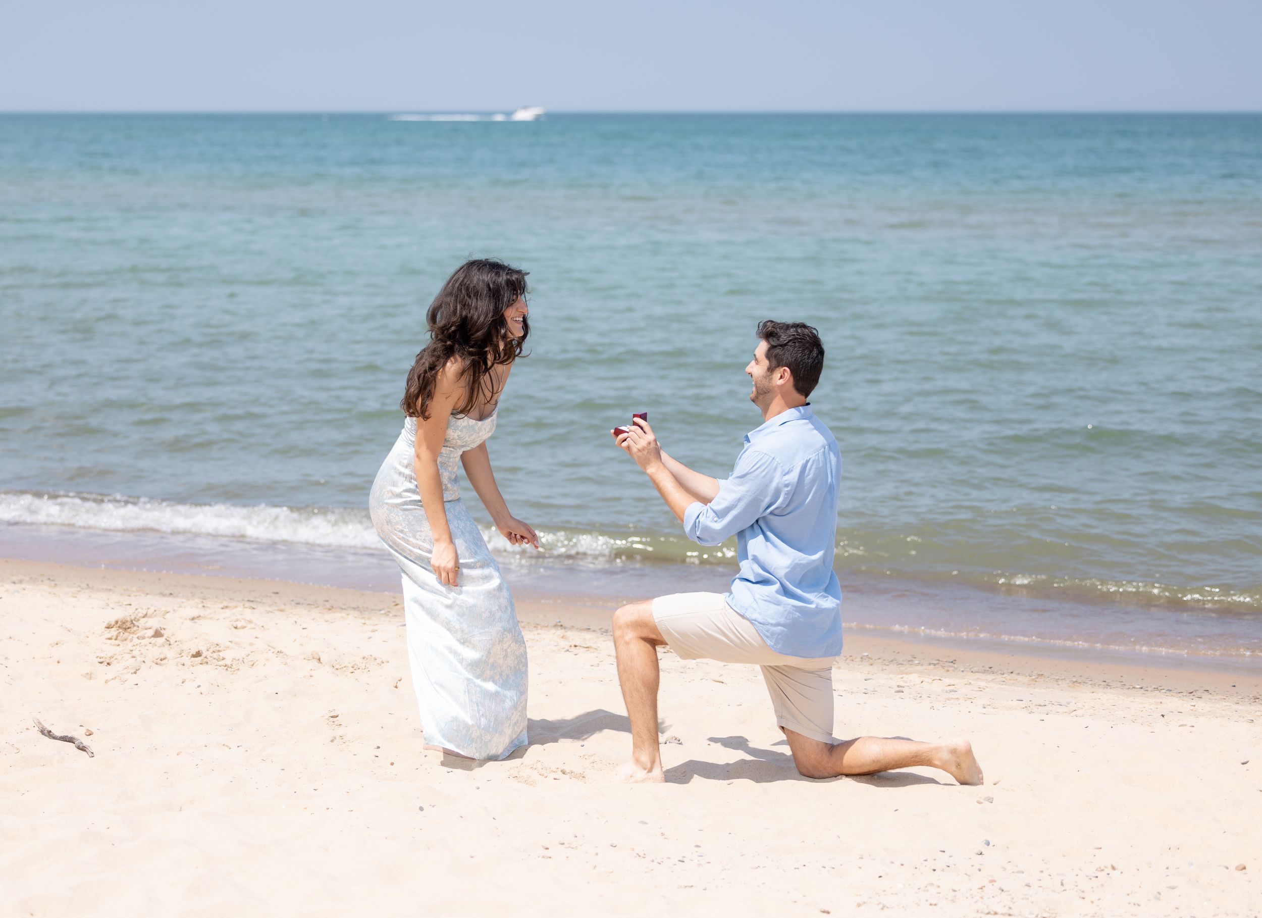 A romantic proposal scene unfolds on a sunny beach with turquoise water and gentle waves in the background.