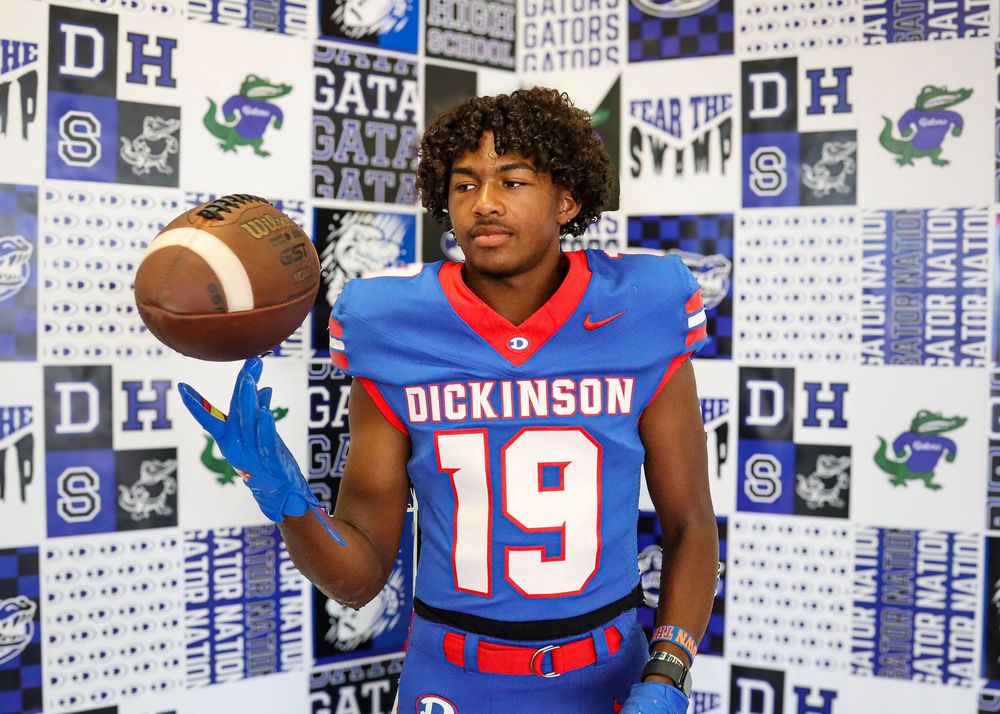 Football player in blue and red uniform poses with ball against backdrop with team logos and graphics.