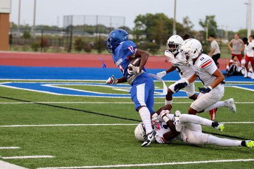 Football player in blue jersey breaks away from defenders on artificial turf field.