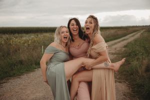 Three bridesmaids laughing together on country road in pastel dresses.