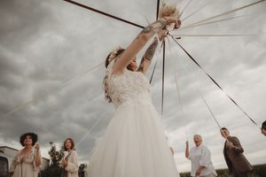 A wedding party gathers outdoors as someone in white reaches upward toward suspended decorations.