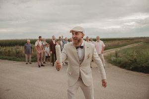Person in light colored suit walks ahead of wedding party group on rural road.