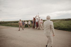 A group celebrates outdoors on a rural path against a cloudy sky wearing neutral-toned wedding attire.