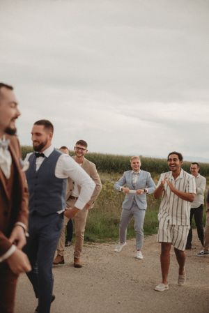 A casual wedding party gathering on a dirt road surrounded by fields shares laughs and smiles.