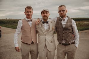 Three groomsmen in coordinating beige and neutral toned suits pose together on a rural path.
