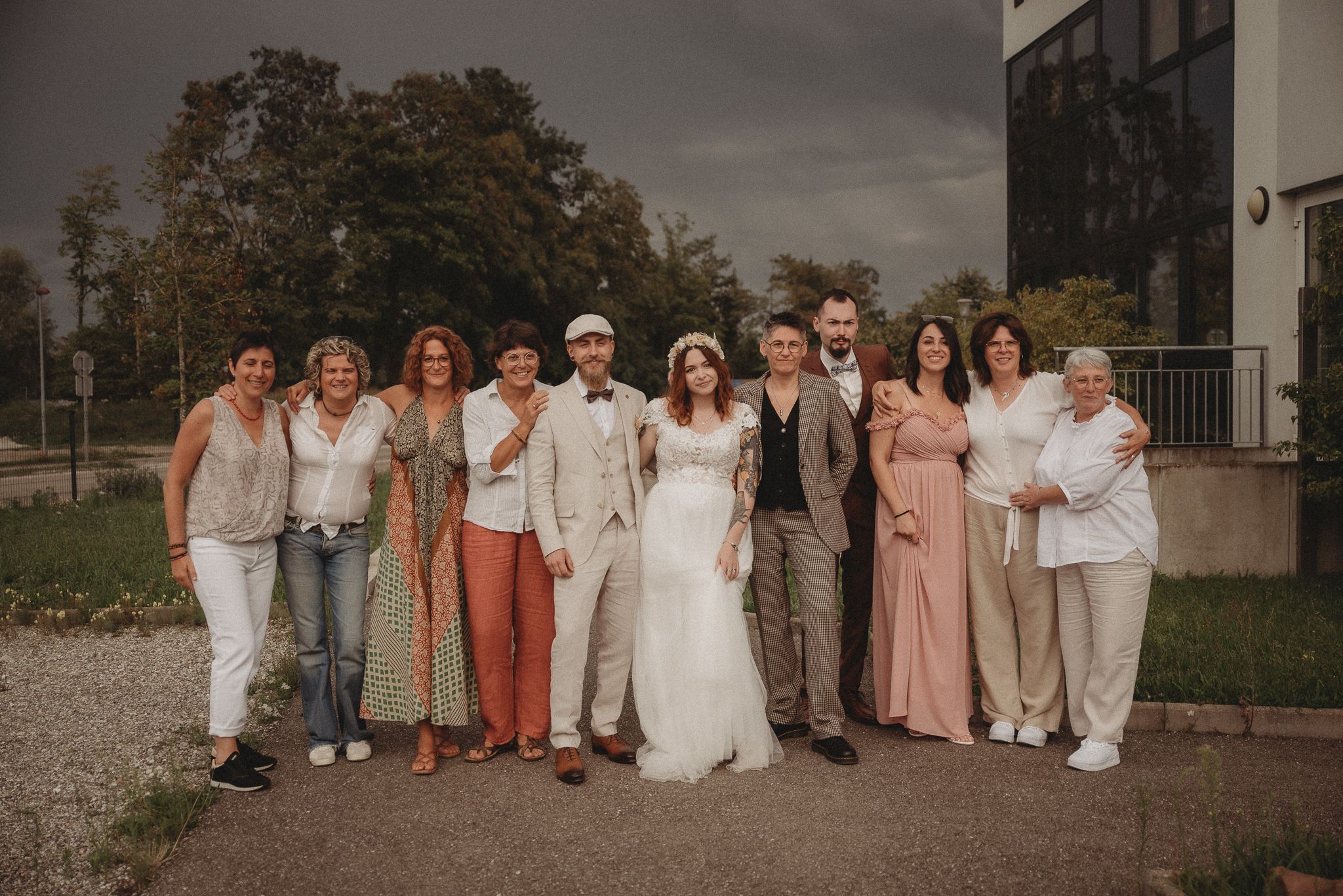 A wedding party group poses outdoors in coordinated neutral and pastel outfits on an overcast day.