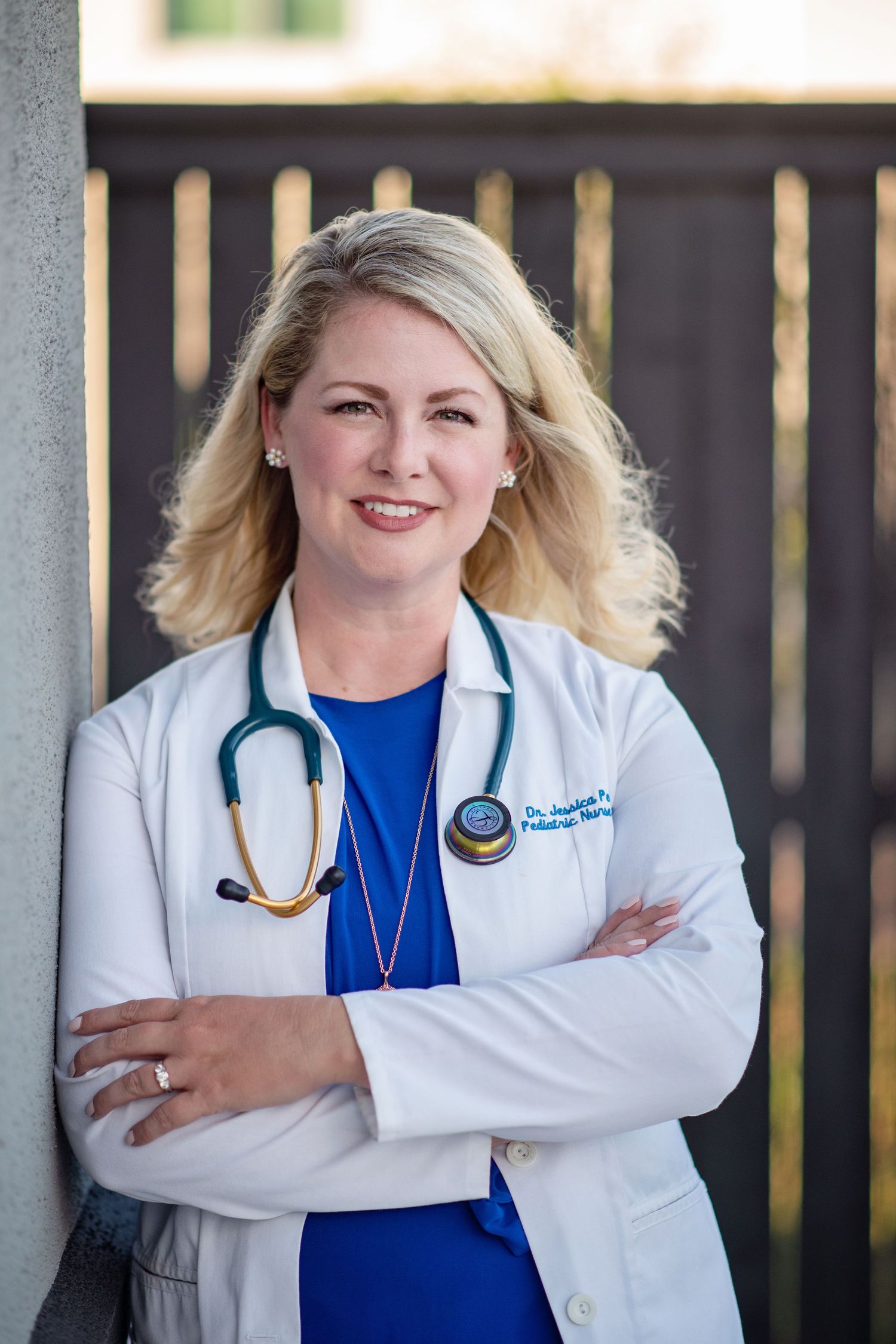 headshot of smiling nurse practitioner leaning against wall wearing white coat and stethoscope