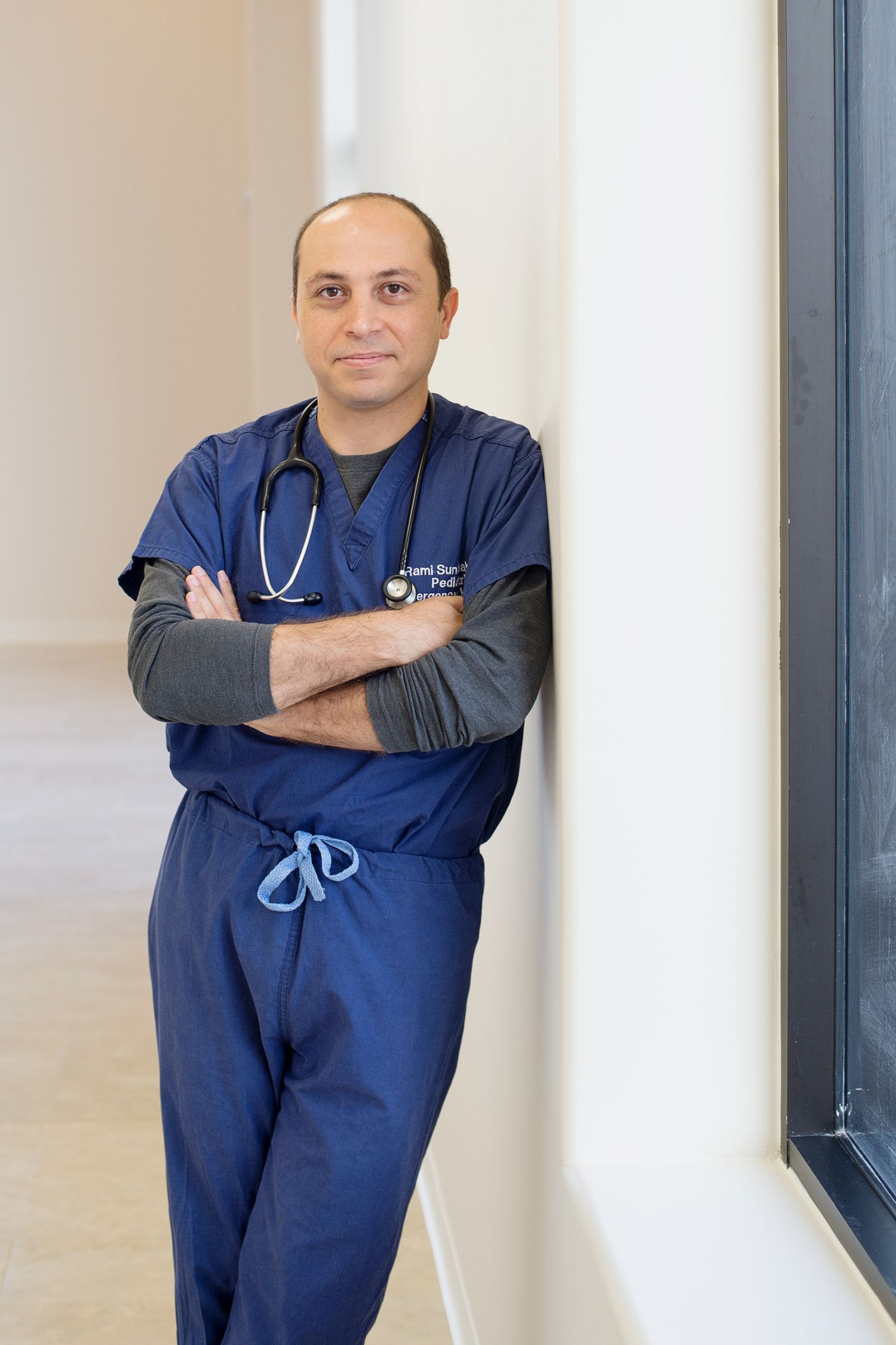 smiling physician wearing scrubs leading against a wall