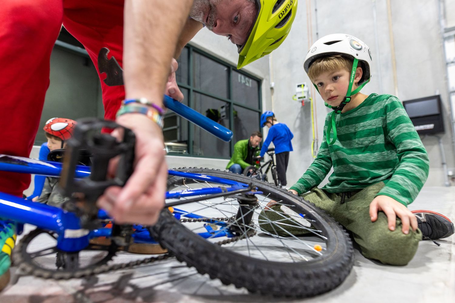 A young boy in a green and white striped shirt is sitting on the ground next to a blue bicycle with a repair person working on it.