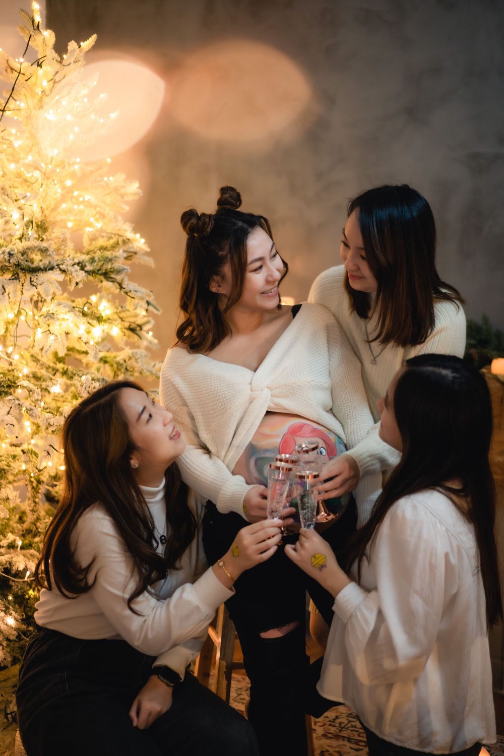 Four friends celebrating together in front of a glowing Christmas tree with white sweaters and holiday cheer.