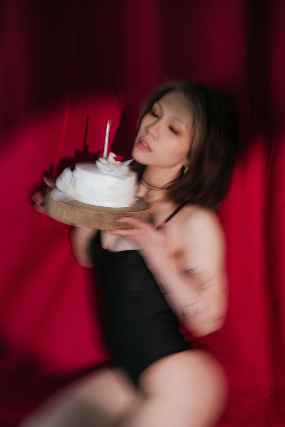 Person in black outfit blowing out candle on white birthday cake against red backdrop.