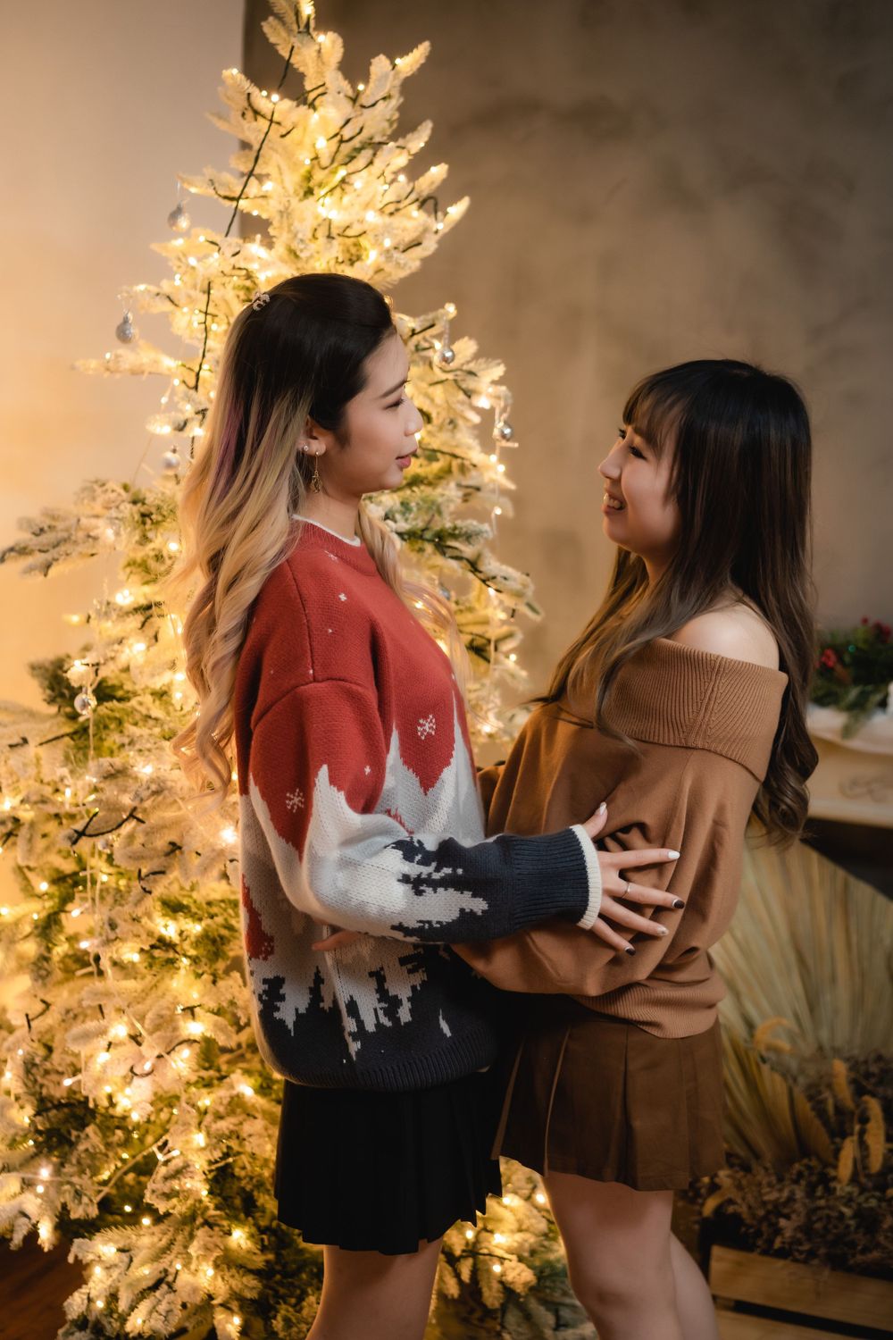 Two friends share an intimate moment in front of a decorated Christmas tree with warm lighting.