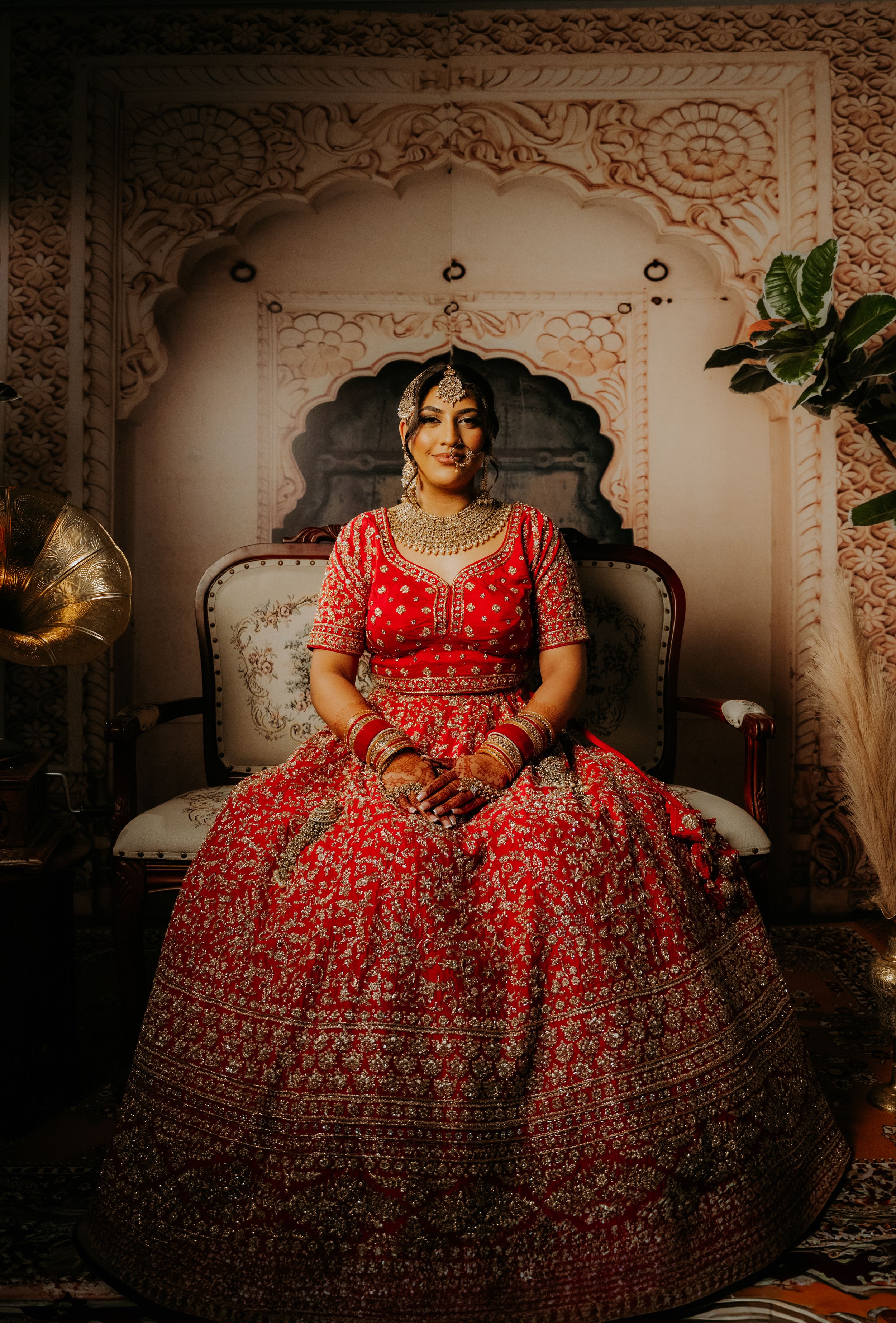 Bride in ornate red lehenga sits on vintage chair against decorative wall background.