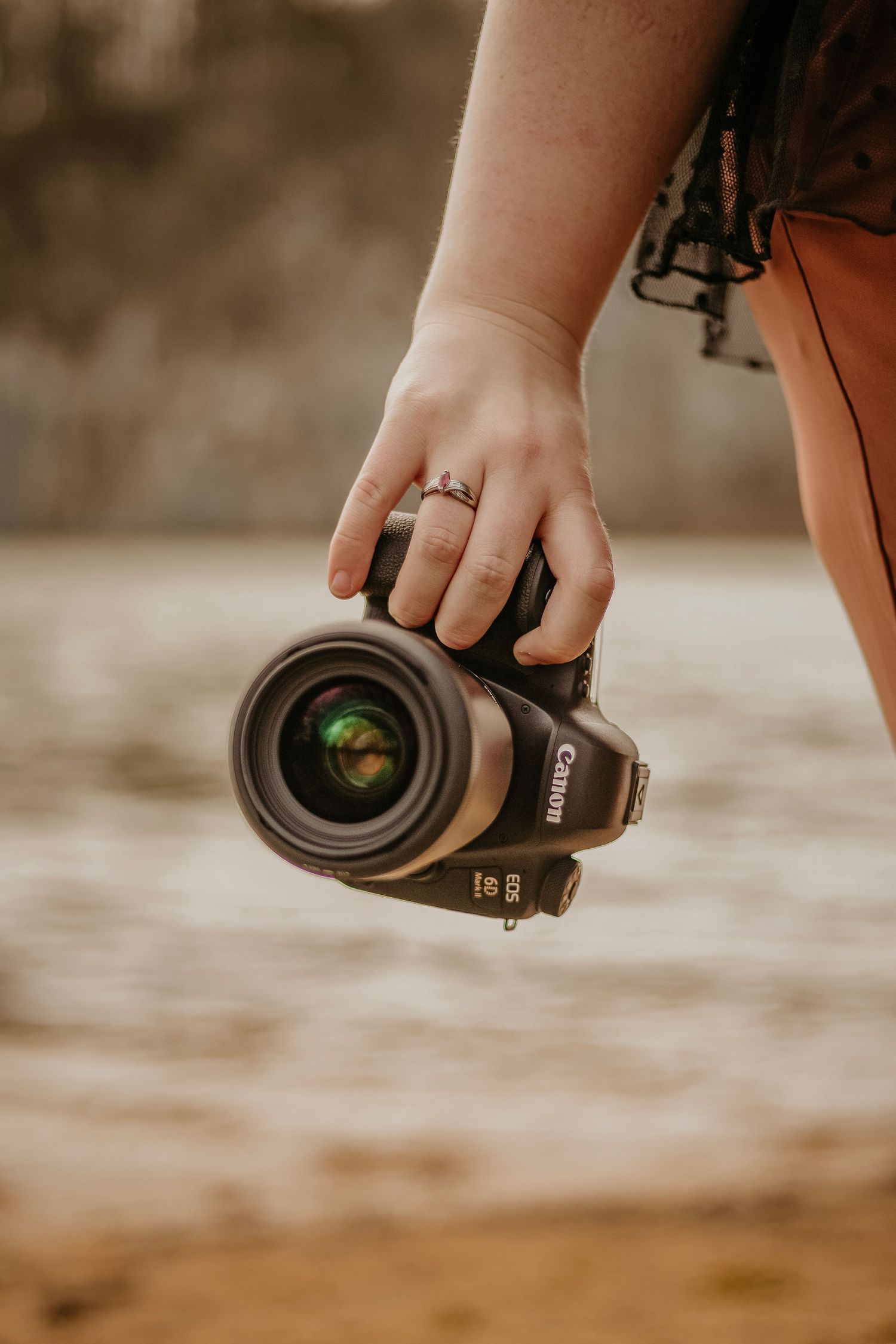 Hand holding a Canon camera with a professional lens against a warm sepia-toned background.