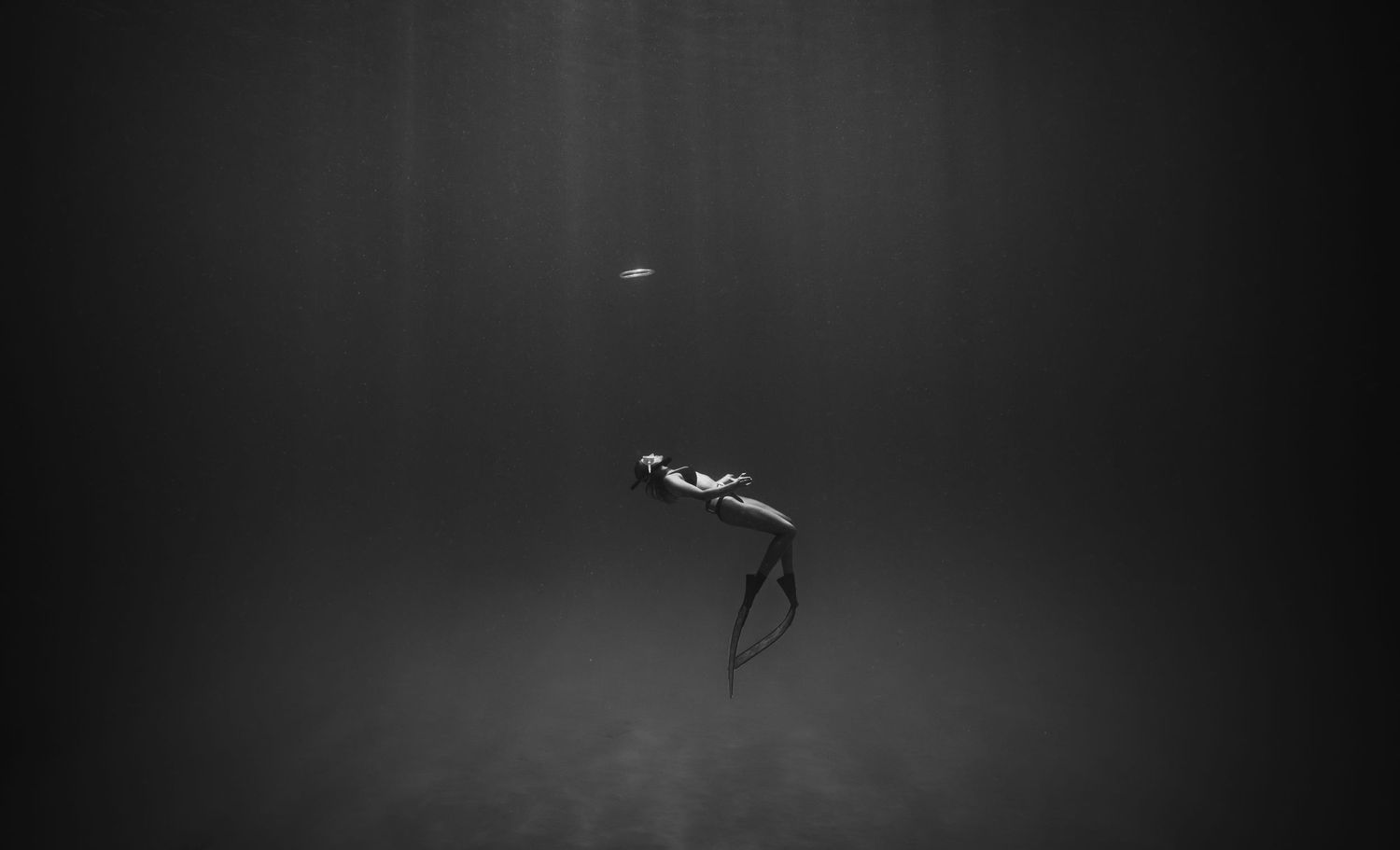 Monochromatic underwater image of a freediver descending into dark ocean depths.