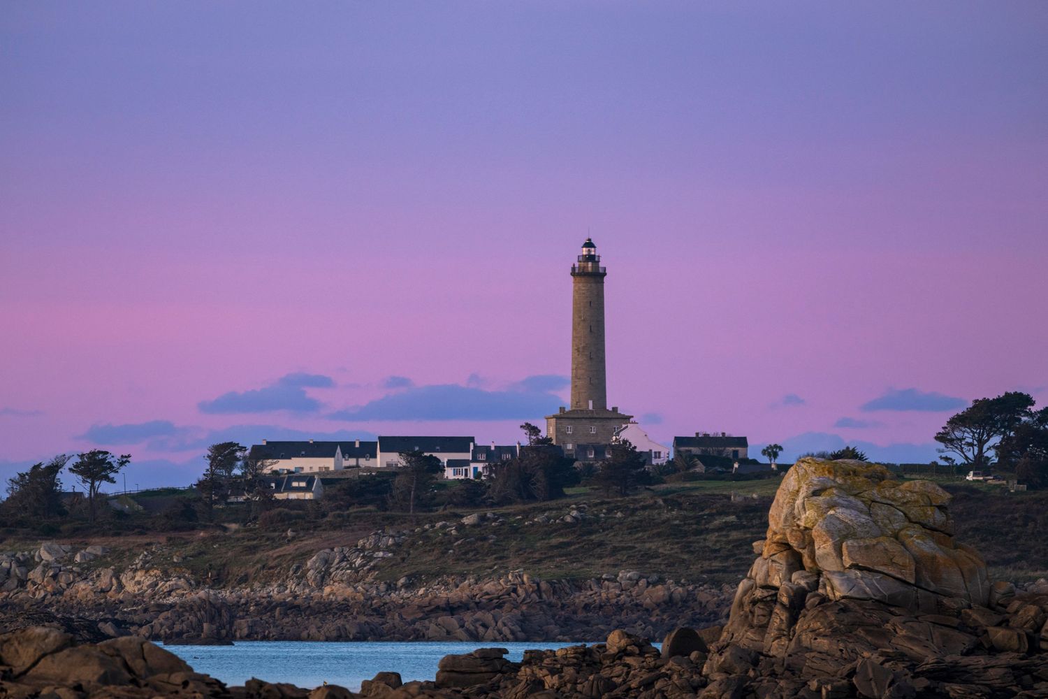 Lighthouse stands majestically on rocky coast during pink sunset with buildings nestled nearby.