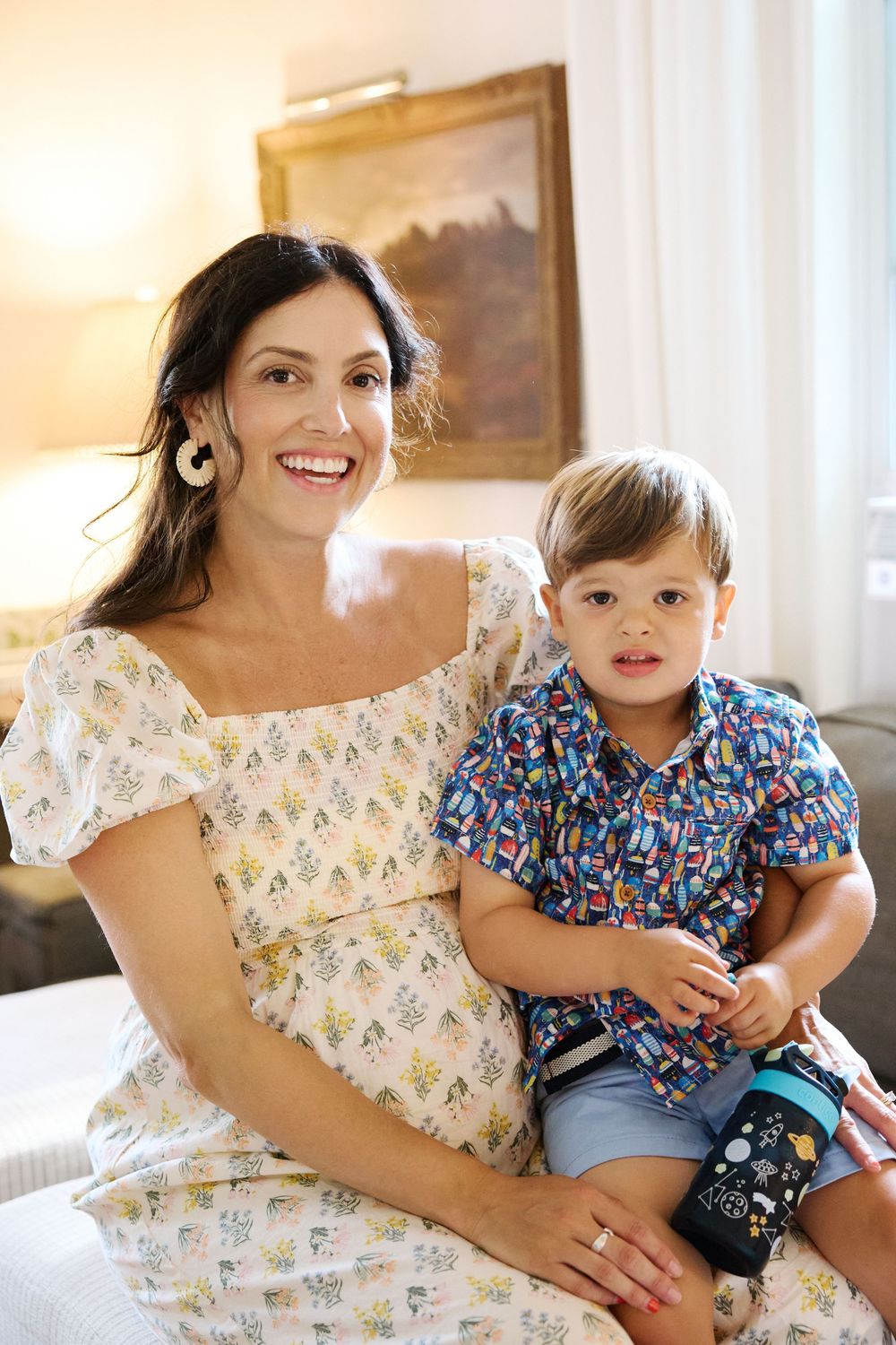 A parent and child sit together on a couch, wearing floral and patterned clothing in a warmly lit room.