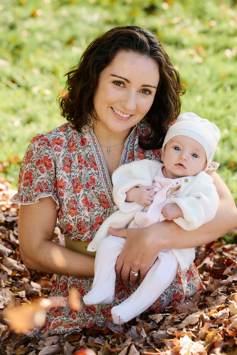 A mother and baby sit together in fallen autumn leaves while wearing floral and white outfits during an outdoor photoshoot.