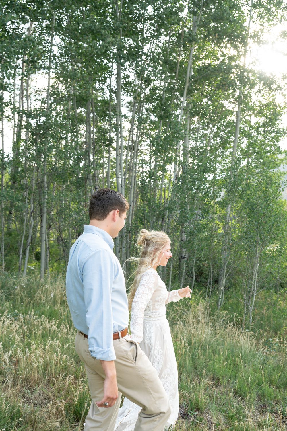 Couple in wedding attire walking through aspen forest, bride in lace gown and groom in light blue shirt.