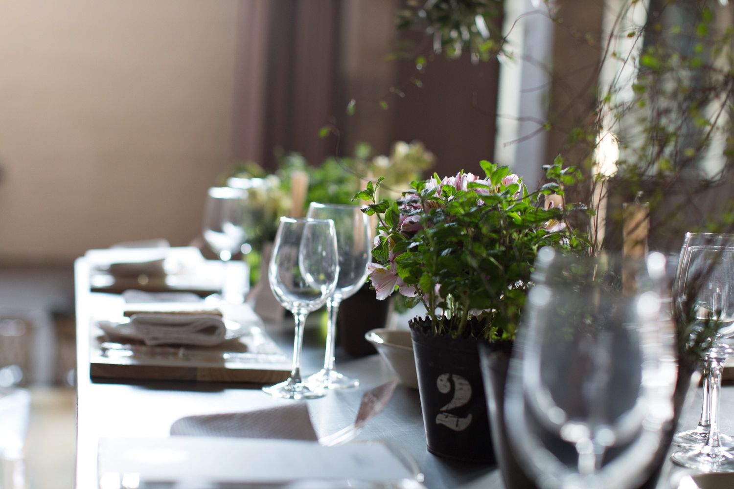 Elegant outdoor table setting with wine glasses, potted flowers, and numbered table markers in sunlight.