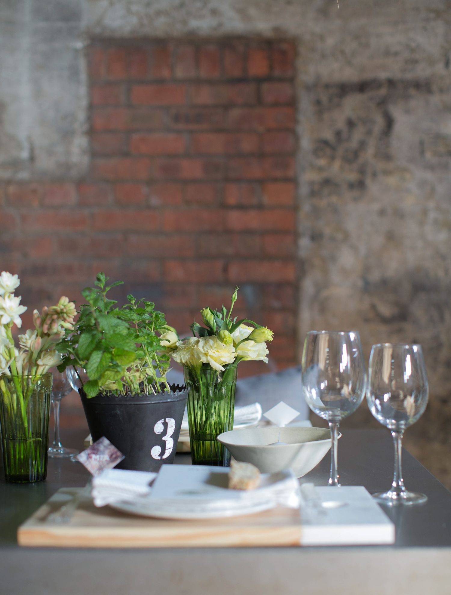 Elegant table setting with white flowers in green vases, wine glasses, and a numbered plant pot centerpiece against brick wall.