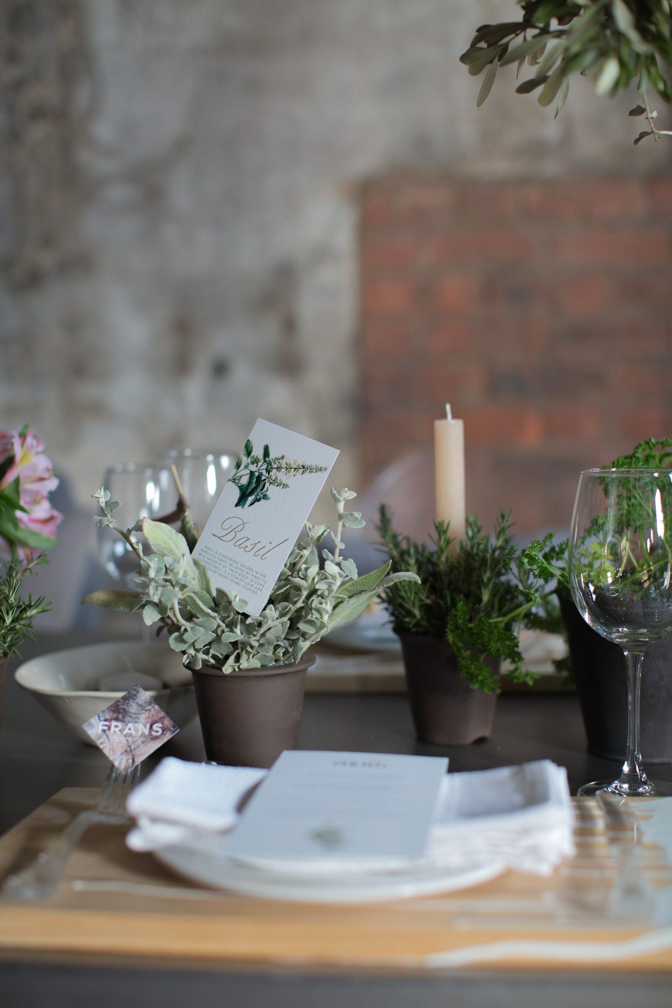 Industrial wedding table setting with potted herb centerpieces, candle, wine glasses, and menu cards on wooden boards.