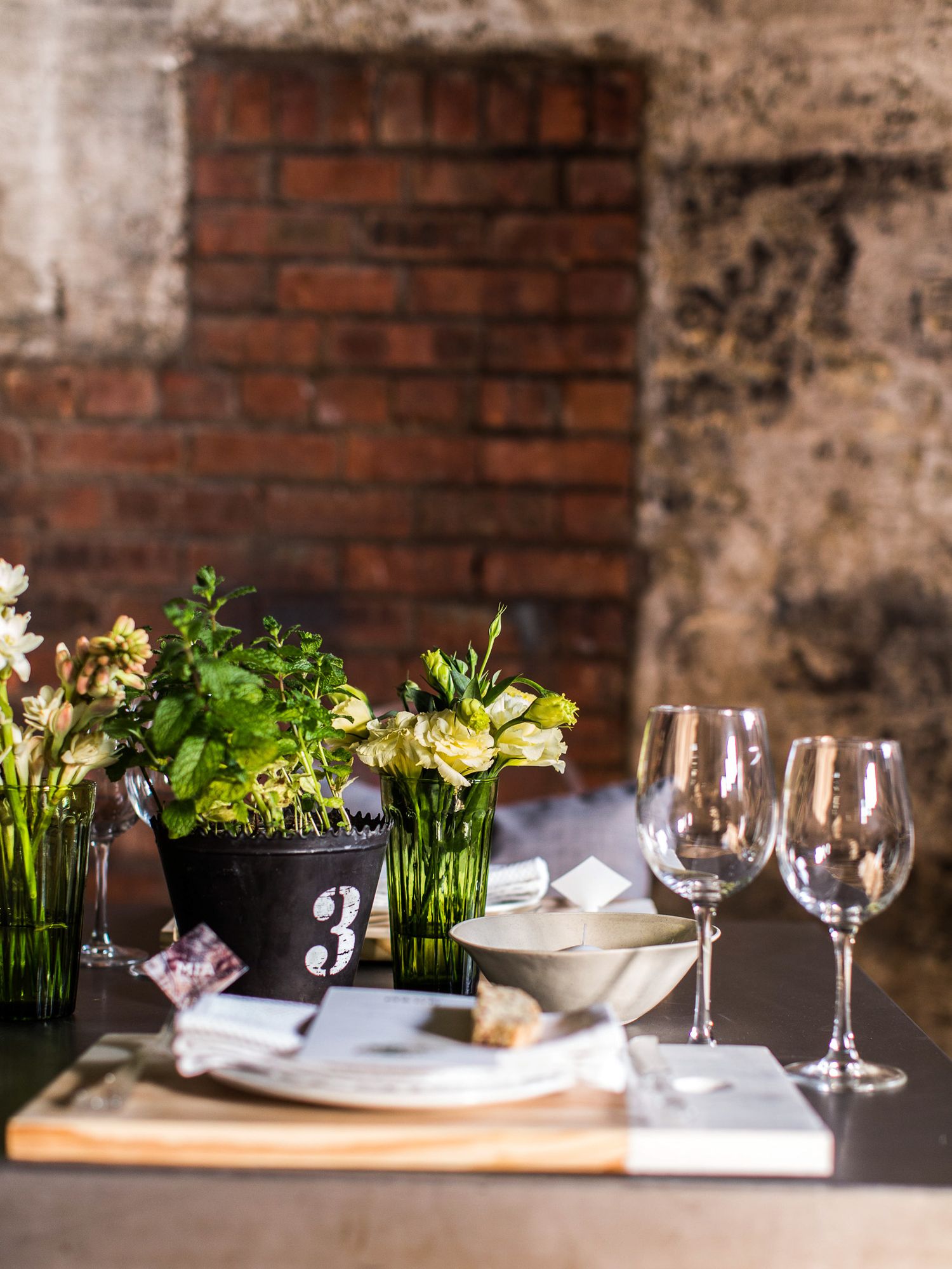 Sophisticated table arrangement with white flowers, wine glasses, and numbered herb pot against exposed brick wall.