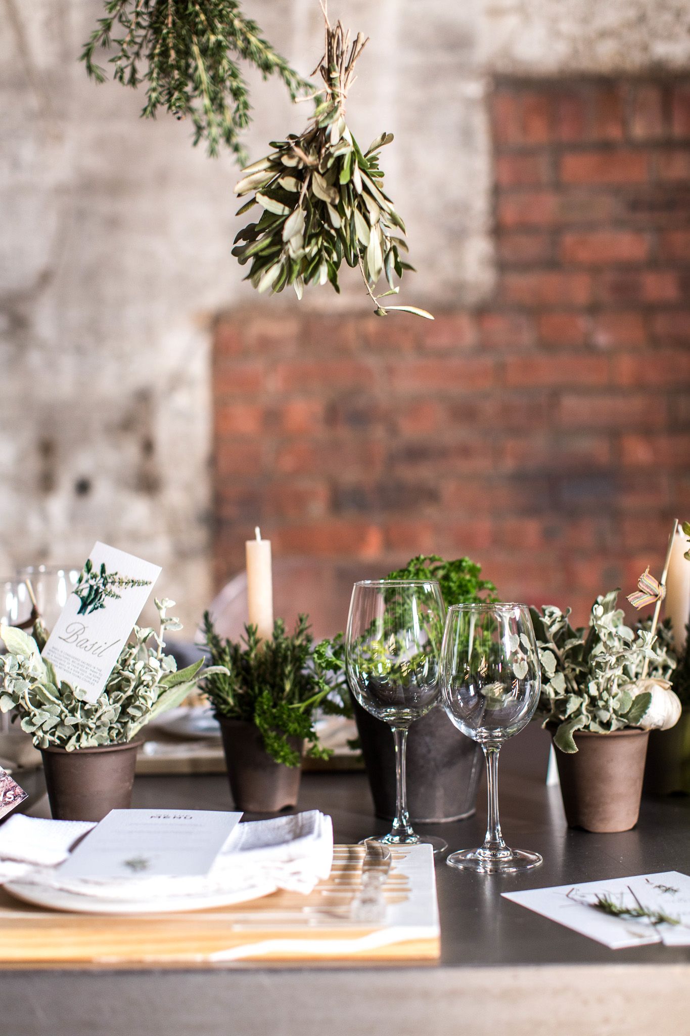 Industrial-chic wedding table decor with hanging herb bundles, potted plants, wine glasses, and brick wall backdrop.