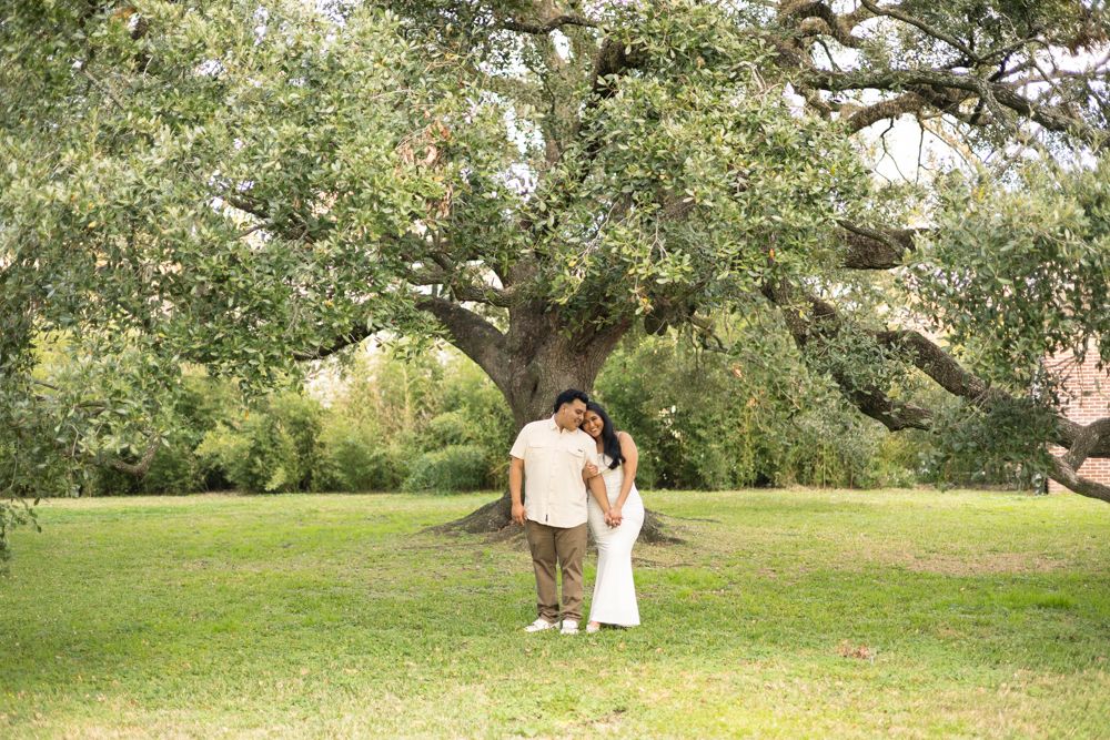 A couple stands together under a large spreading tree in a lush green park setting.