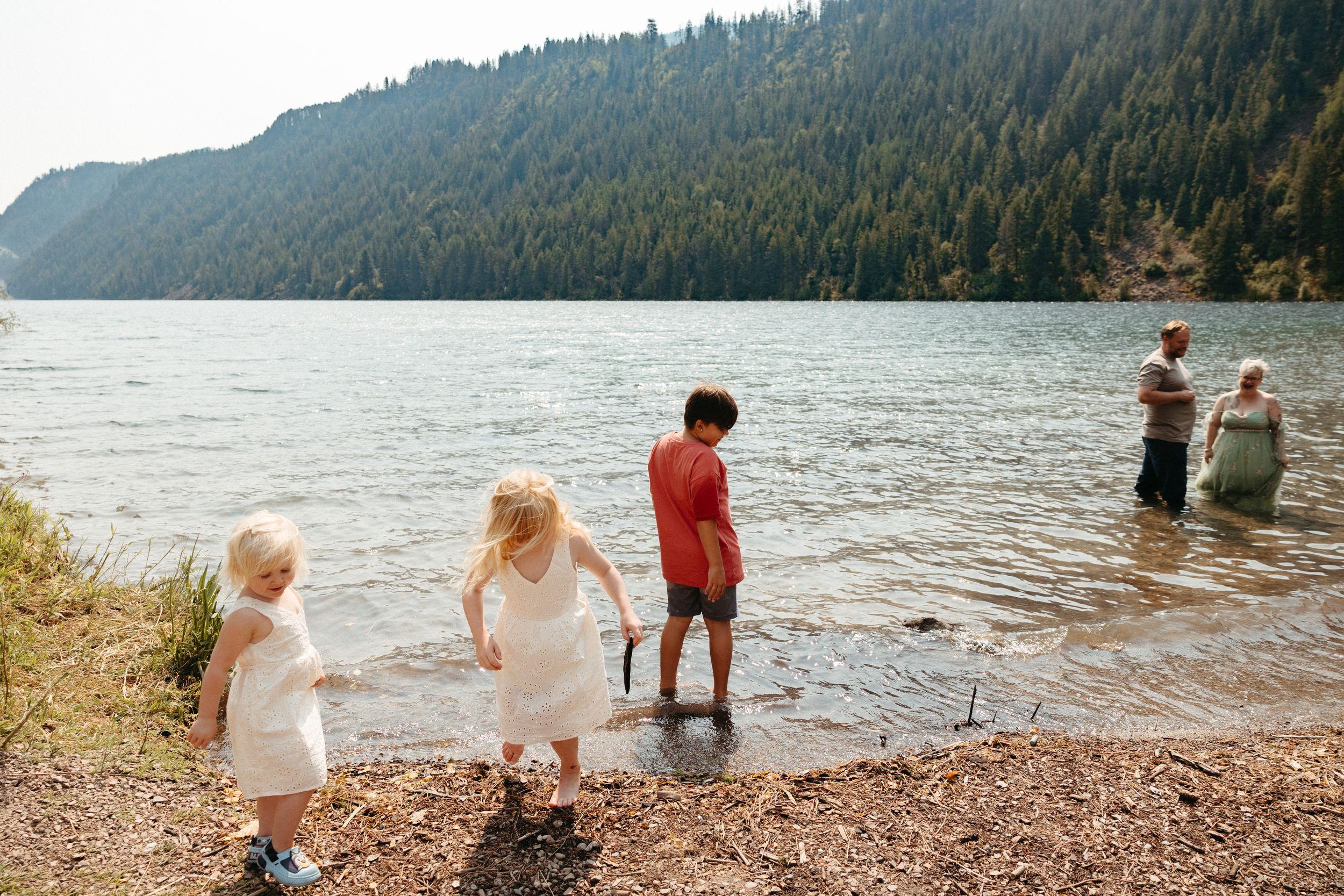 Children playing at the edge of a calm mountain lake surrounded by dense evergreen forest on a sunny day.