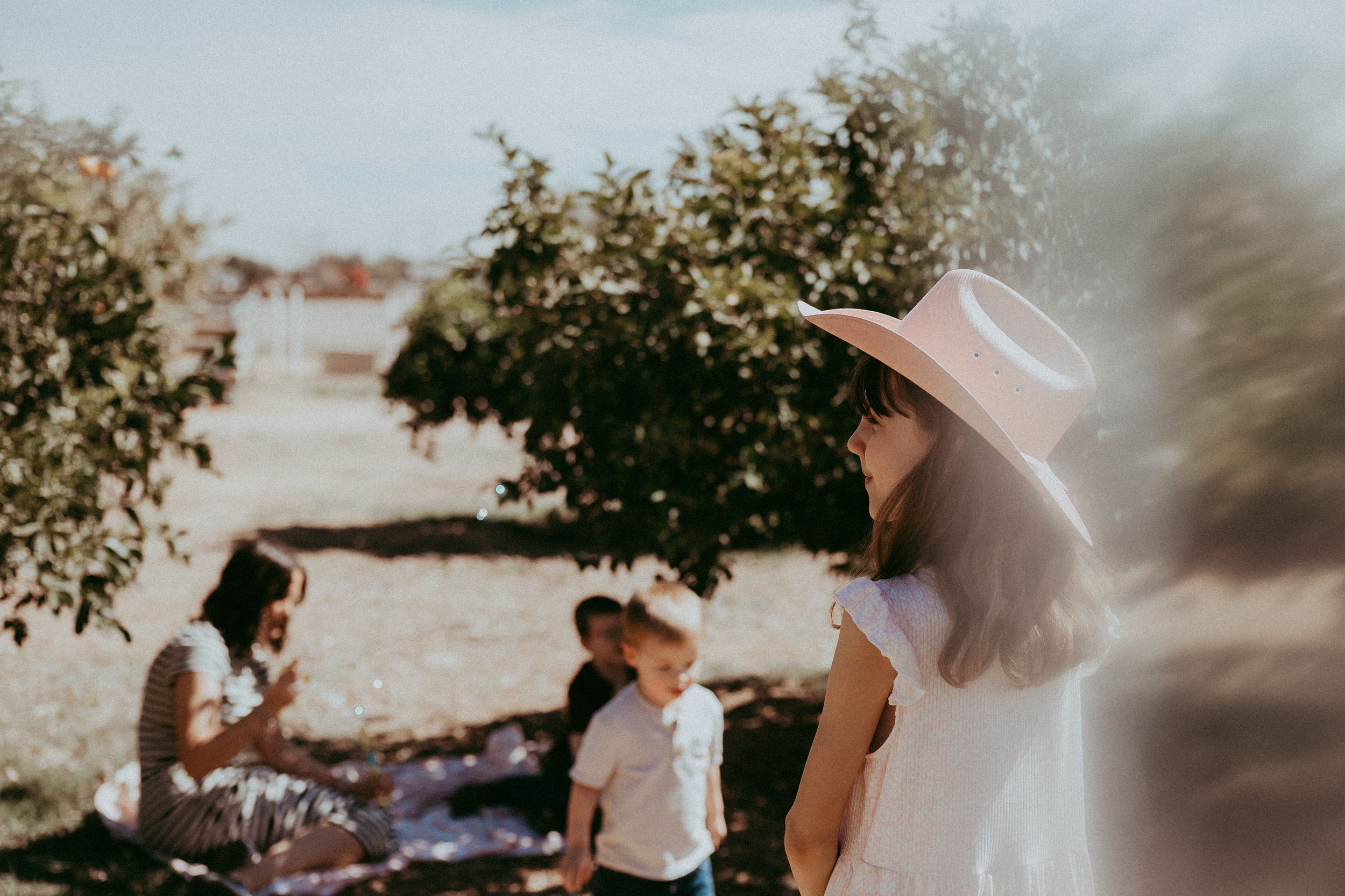 Little girl in white dress and hat stands outdoors near trees while family enjoys a picnic in the background.