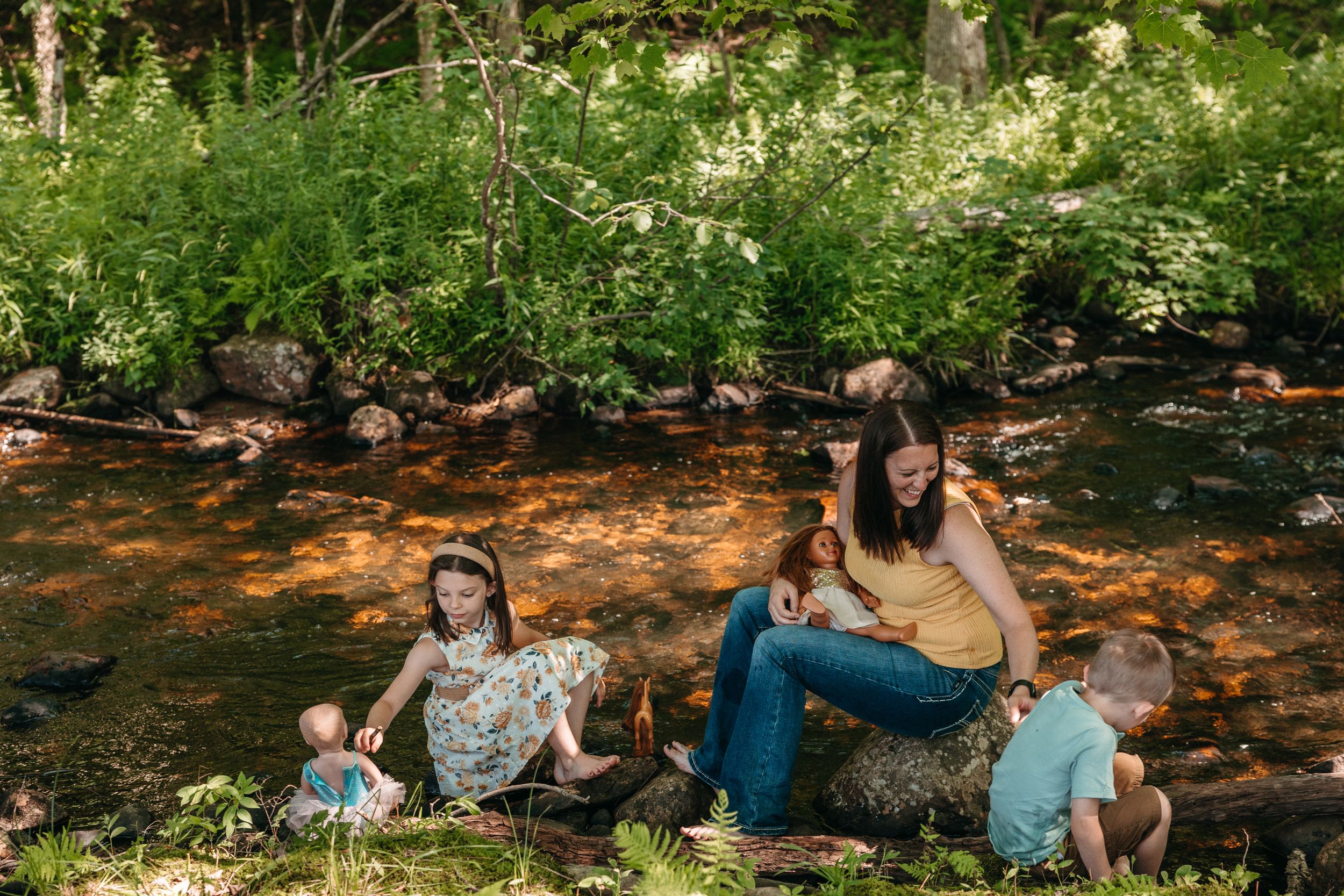 Family relaxing by a sunlit forest creek, mother with children playing on rocks surrounded by lush green trees.