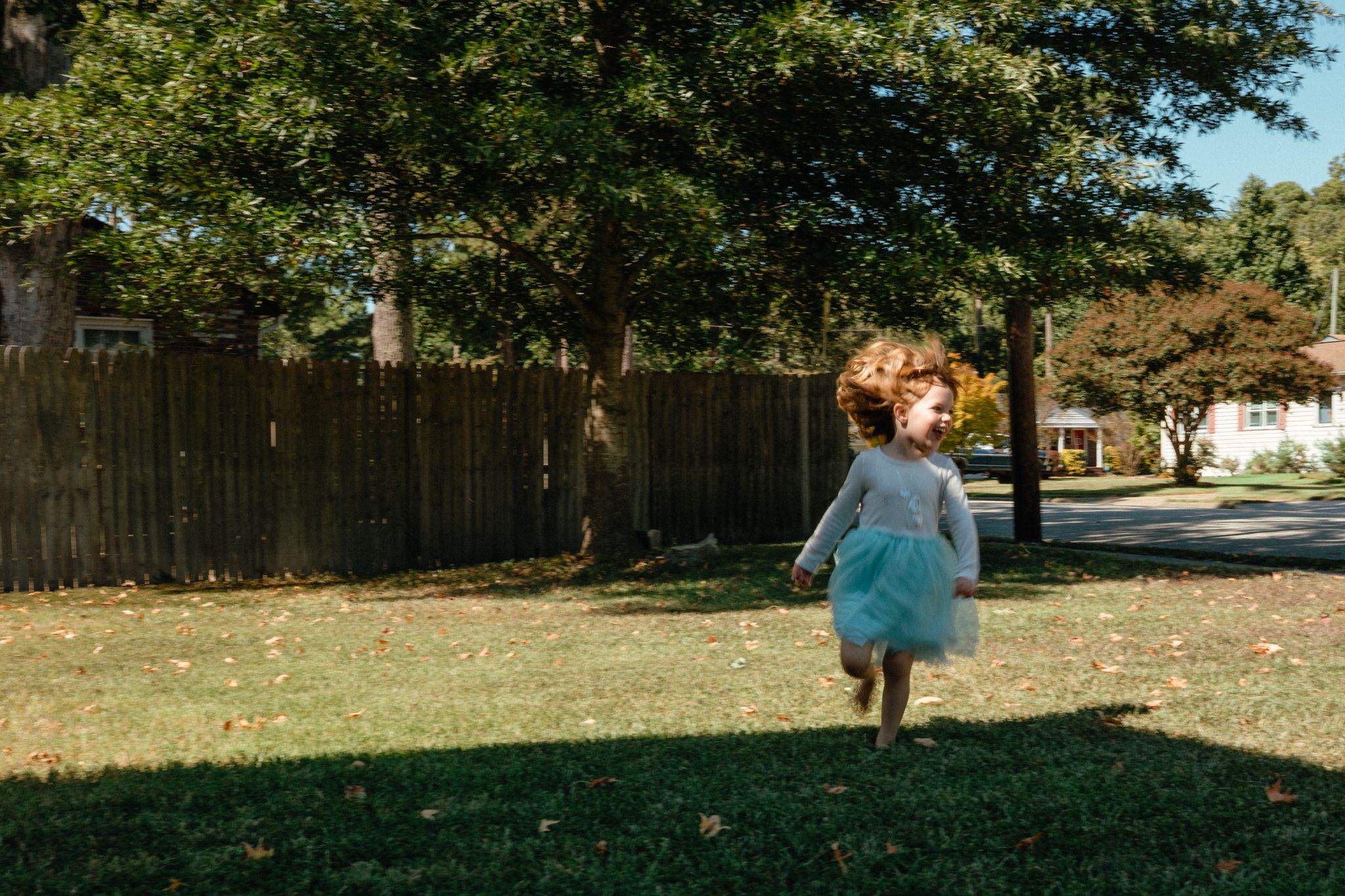 A young girl in a blue tutu dress runs across a green lawn near a wooden fence and large trees on a sunny day. Whitney Popp, Phoenix Arizona