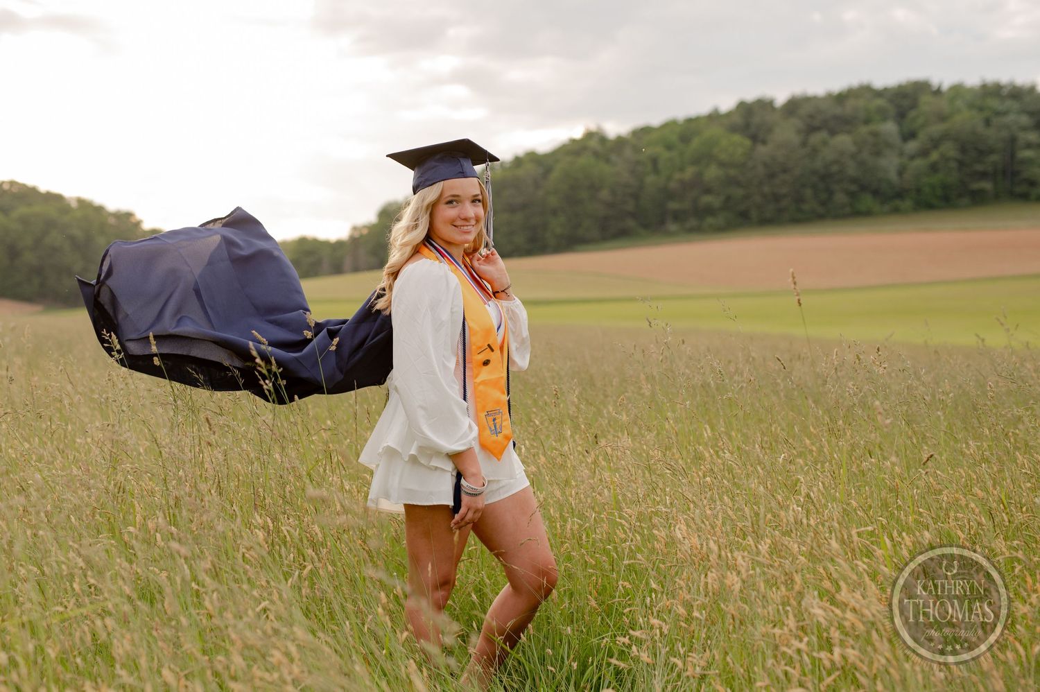 Graduate in flowing gown holds cap while walking through tall grass field at sunset.