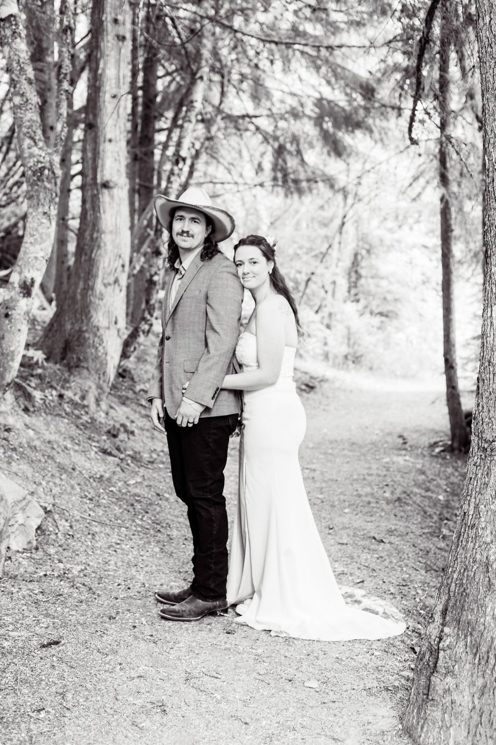 A romantic black and white wedding photo of a couple standing back-to-back on a forest path surrounded by tall trees.