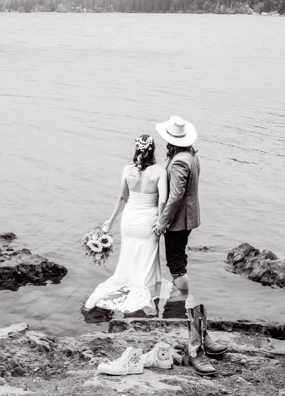Black and white wedding photo showing a couple standing on rocky shoreline with boots in foreground beside a lake.