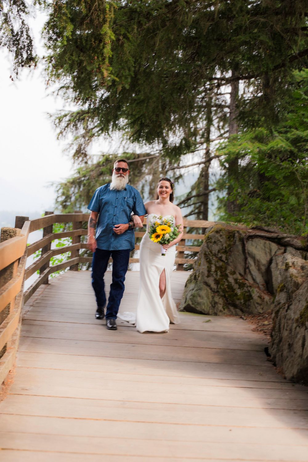A couple walks along a scenic wooden boardwalk surrounded by evergreen trees at a coastal location.