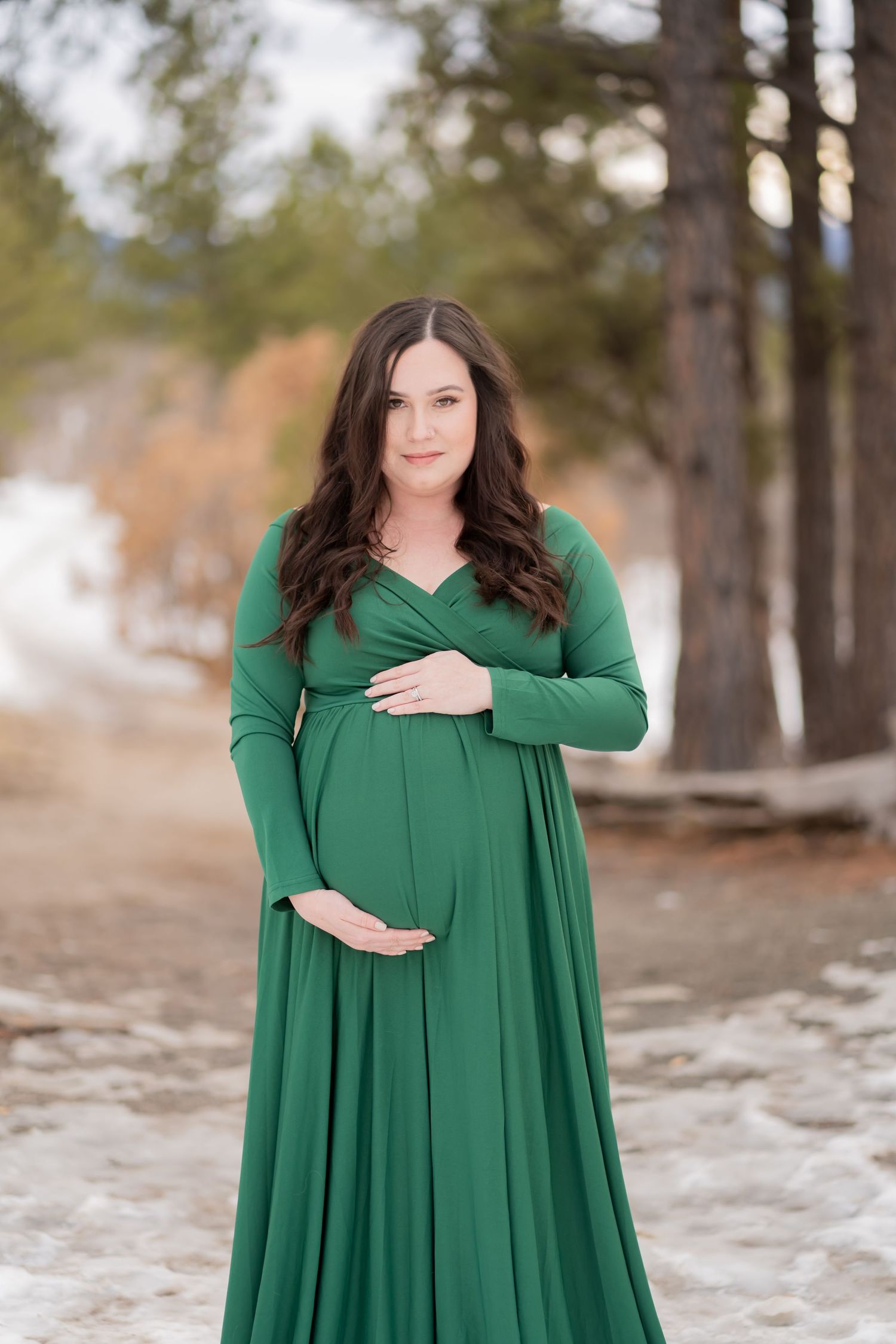 Elegant maternity portrait in emerald green gown against snowy winter forest backdrop.