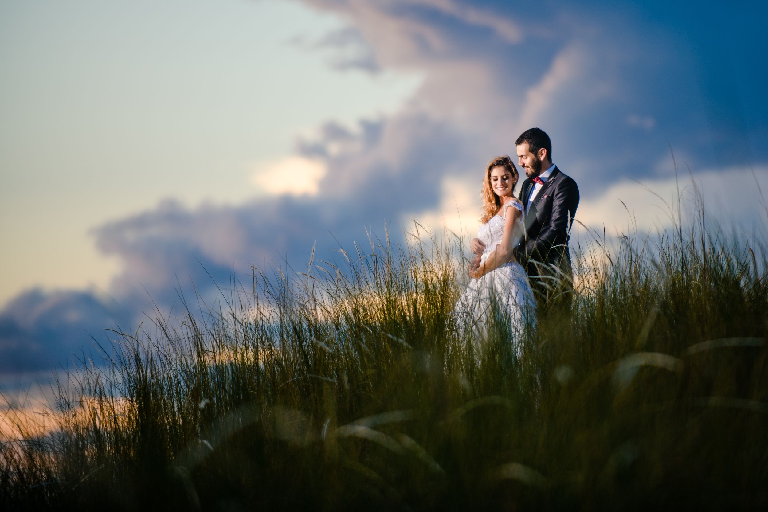 Newlywed couple embraces in a serene grassy field at sunset, with a dramatic sky creating a romantic and timeless wedding moment.