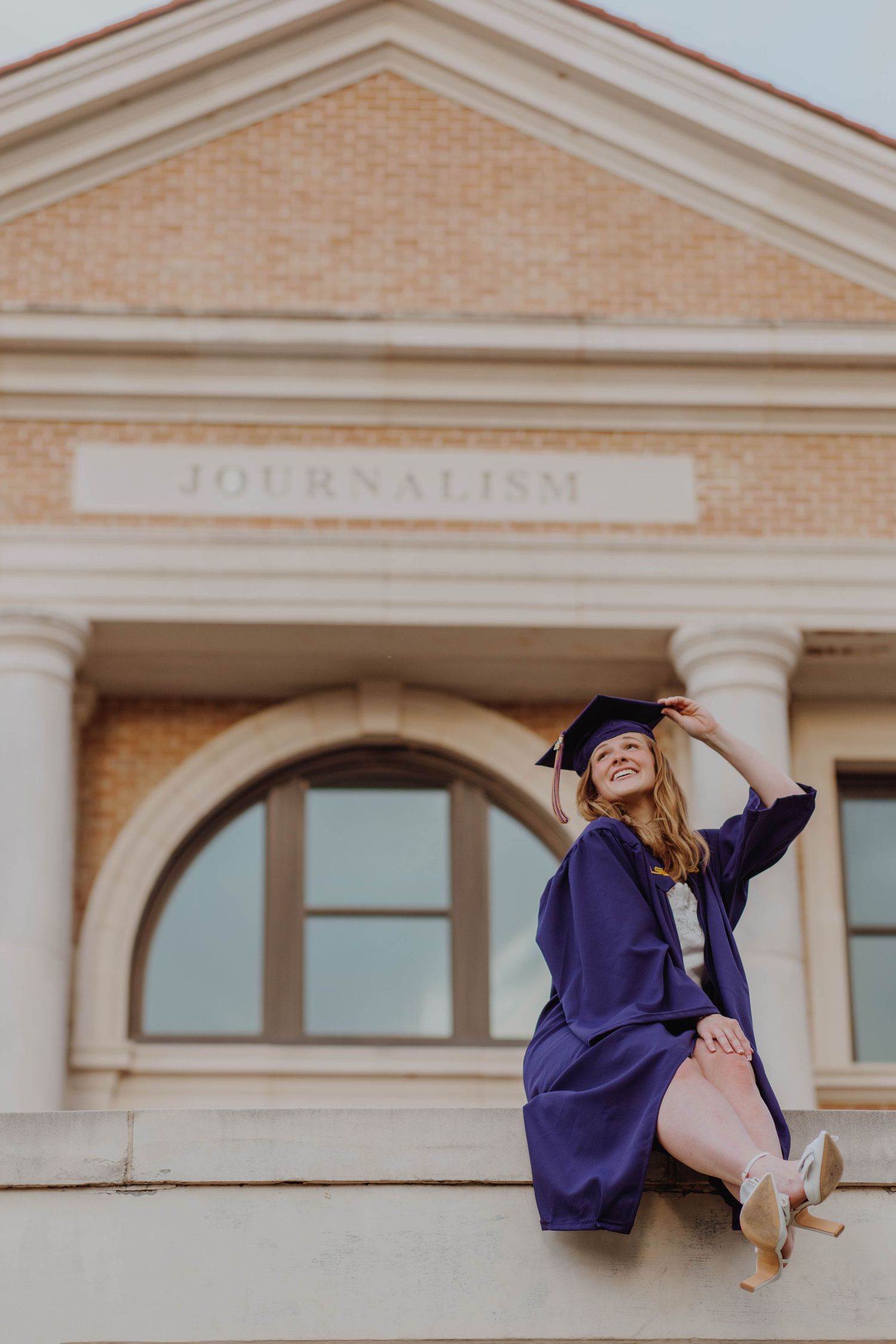 Graduate in purple cap and gown poses playfully outside journalism building.