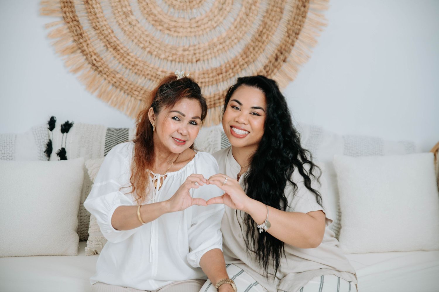 Two people make a heart shape with their hands while sitting on a white couch.