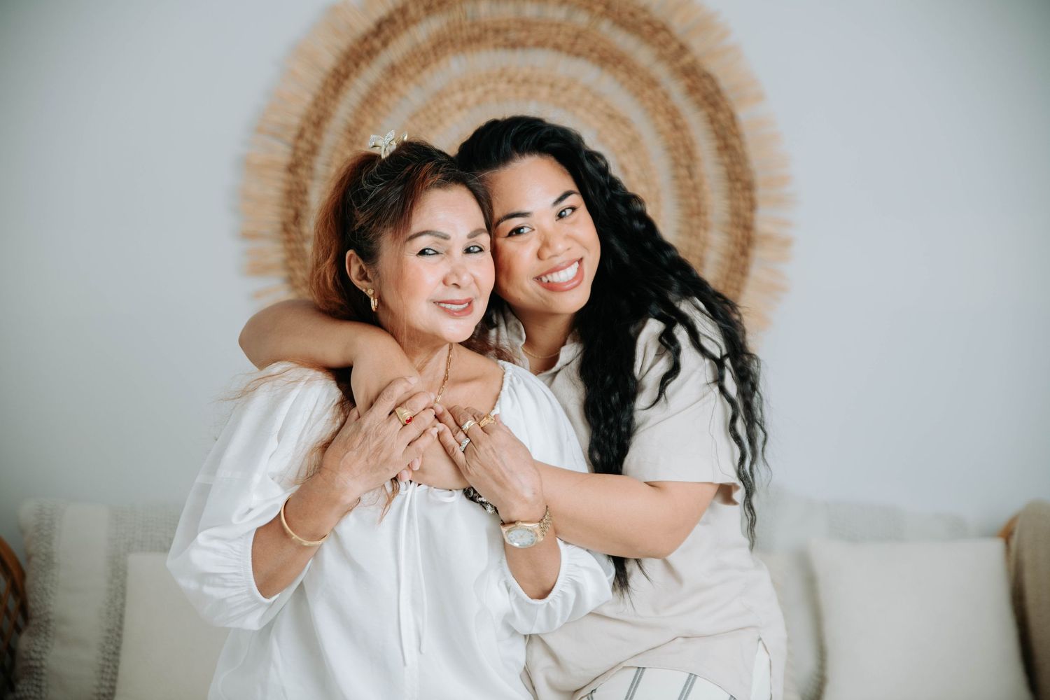 Two people embrace while sitting on a couch with a bohemian circular wall hanging in the background.
