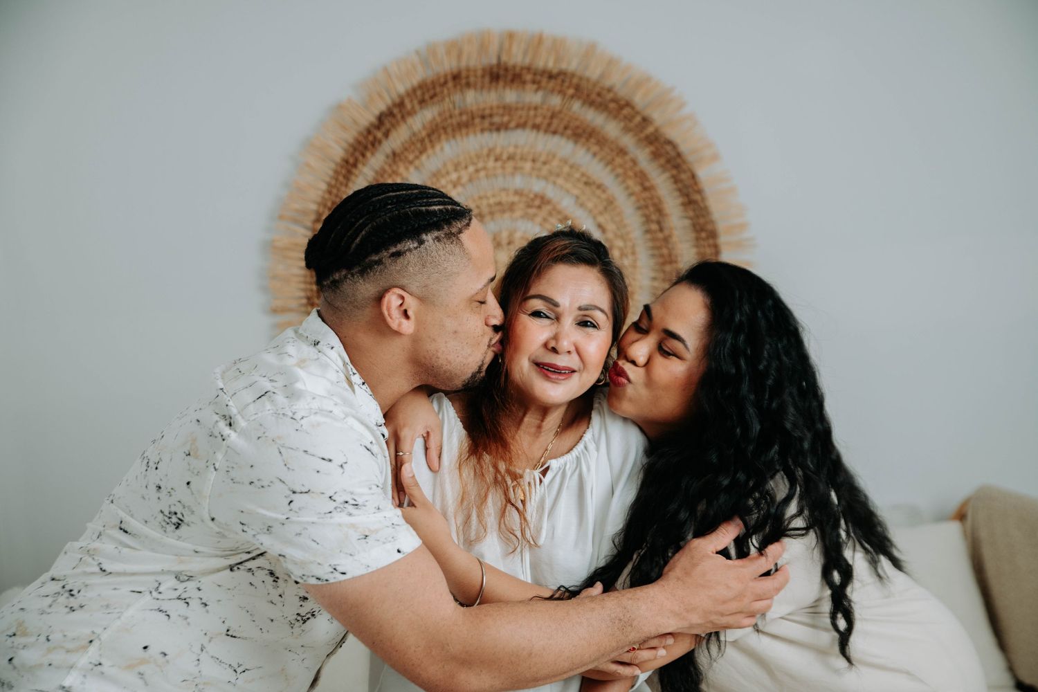 Three people share an affectionate moment on a couch with a decorative circular wall hanging behind them.