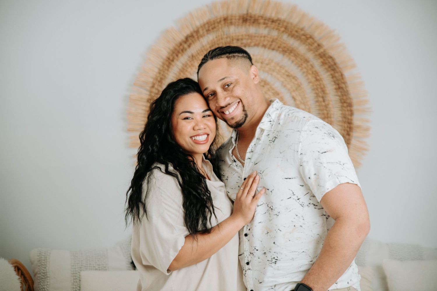 A couple poses together against a woven circular wall hanging in matching white outfits.